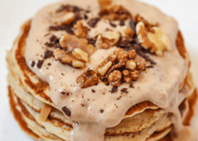 A stack of chunky monkey pancakes on a plate, topped with peanut butter sauce, walnuts and choc chips