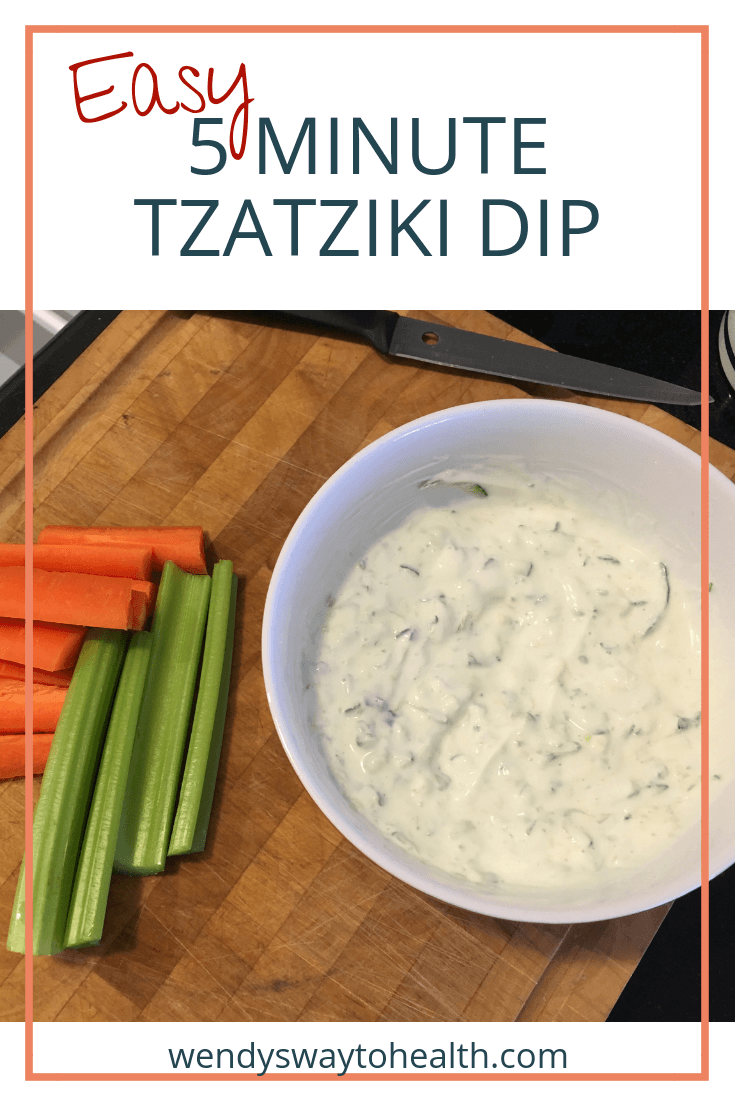 a bowl of tzatziki dip sitting on a chopping board beside carrot and celery sticks
